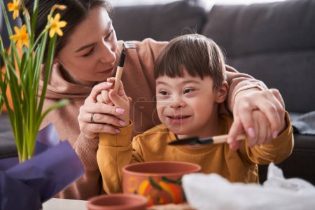 Téléchargez les photos : Mère caucasienne enseignant à son petit enfant atteint du syndrome du duvet à remplacer les fleurs dans les pots à la maison. Garçon se sentir heureux - en image libre de droit