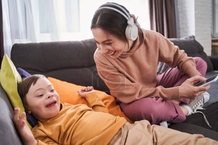 Téléchargez les photos : Femme gaie tenant smartphone et riant à haute voix à son fils avec des troubles génétiques tout en se relaxant sur le canapé ensemble - en image libre de droit
