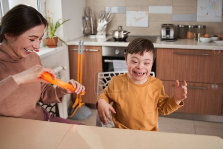 Téléchargez les photos : Je suis heureuse. Petit garçon avec des troubles génétiques riant à haute voix tandis que sa mère soufflant des bulles de savon avec lui à la cuisine. Photo de stock - en image libre de droit