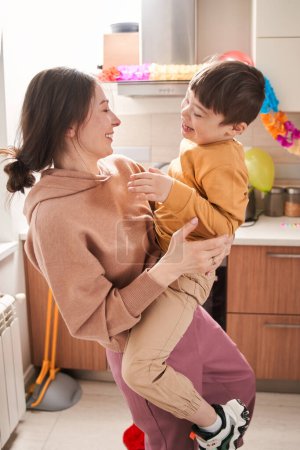 Téléchargez les photos : Vue verticale de la femme ravie tenant par les mains son enfant mâle avec un trouble génétique tout en s'amusant à la cuisine - en image libre de droit