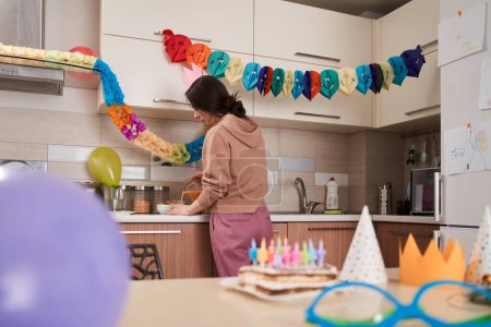 Téléchargez les photos : Gâteau d'anniversaire avec des bougies debout à la table avec d'autres décorations de fête à proximité. Mère debout à la cuisine à l'arrière-plan - en image libre de droit