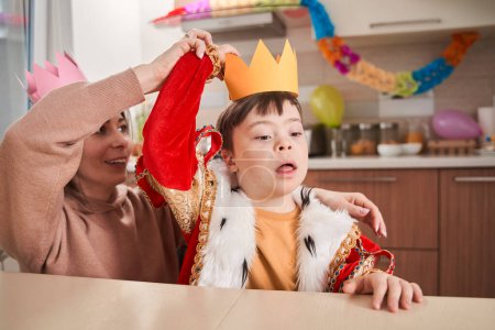 Téléchargez les photos : Petit garçon avec trouble génétique portant costume de roi se préparant à la fête avec sa mère alors qu'il était assis à la table à la cuisine. Concept de famille - en image libre de droit