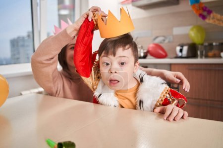 Téléchargez les photos : Garçon mignon avec trouble génétique portant costume roi assis à la table et se préparant à la fête avec sa mère. Concept de famille - en image libre de droit
