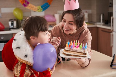 Téléchargez les photos : Petit garçon avec le syndrome de Down regardant loin tandis que sa mère ravie debout avec gâteau d'anniversaire aux mains. Enfant préparant des bougies soufflantes - en image libre de droit