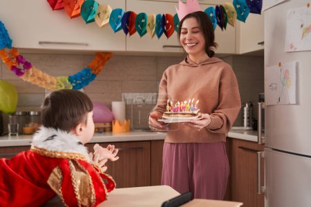Téléchargez les photos : Petit garçon avec trisomie 21 regardant sa mère ravie avec un gâteau d'anniversaire aux mains. Enfant préparant des bougies soufflantes - en image libre de droit