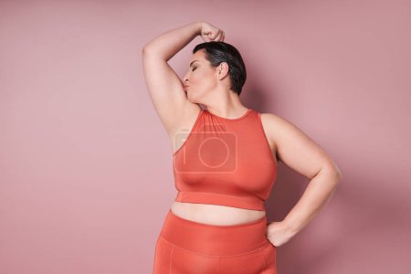 Young caucasian plus size female model kissing biceps after training on pink background. Concept of sport, healthy lifestyle, body positive, equality