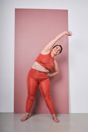 Foto de cuerpo entero de mujer gorda sonriente con sobrepeso usando ropa deportiva haciendo ejercicio en forma aislada en el fondo rosa del estudio. Ejercicio deportivo, fitness y cuerpo concepto positivo