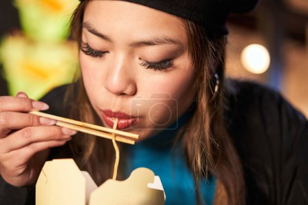 Photo for Focused girl eating noodles outdoors in neon illuminated urban street. Hipster female in trendy clothes enjoying nightlife in city - Royalty Free Image