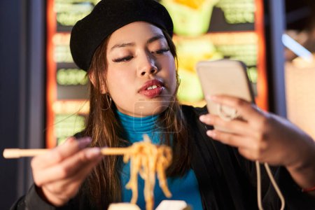 Photo for Focused girl eating noodles and using mobile phone in neon illuminated urban street. Hipster female in trendy clothes enjoying nightlife in city - Royalty Free Image