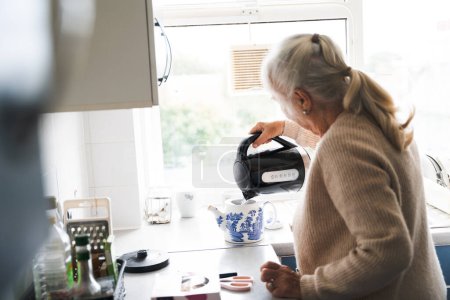 Téléchargez les photos : Une femme âgée verse le thé d'une théière dans la cuisine sur fond de fenêtre - en image libre de droit