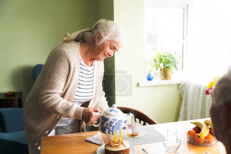 Téléchargez les photos : Une femme âgée verse le thé d'une théière dans la salle à manger pour son mari. Anniversaire célébrer - en image libre de droit
