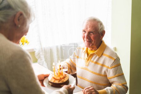 Foto de La esposa trajo un pastel con velas a su anciano esposo, y el celebrante del cumpleaños está listo para pedir deseos y soplar las velas. - Imagen libre de derechos