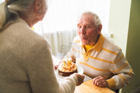 An elderly man is blowing out the candles on his celebratory cake and making a wish.