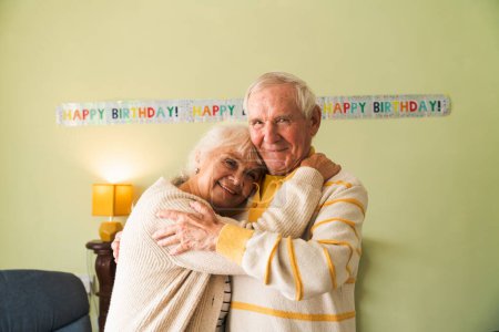 Photo for Happy seniors embrace each other against a birthday celebration backdrop. Concept of elderly longevity at home. - Royalty Free Image