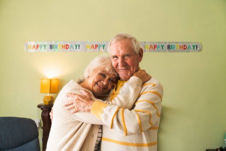 Photo for Happy seniors embrace each other against a birthday celebration backdrop. Concept of elderly longevity at home. Looking to the camera together. - Royalty Free Image