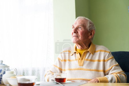 Photo for The happy an elderly man, sits at the table with a tea in the background. Looking to the future and dreaming - Royalty Free Image