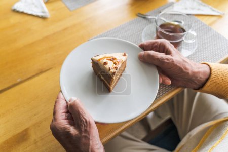 Photo for Close-up of an elderly man holding a plate with a cake against the background of a table. - Royalty Free Image