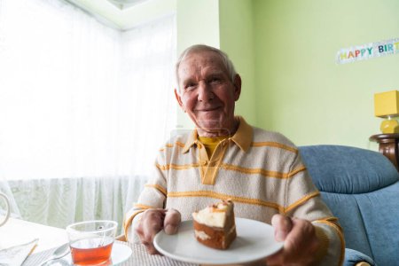 Photo for The happy birthday celebrant, an elderly man, sits at the festive table with a cake and tea in the background. - Royalty Free Image