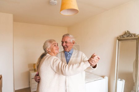 Téléchargez les photos : Un couple de personnes âgées dansant ensemble dans leur chambre bien éclairée, années heureuses ensemble. Concept de vieillissement. - en image libre de droit