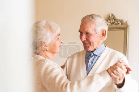 Photo for An elderly couple dancing together in their well-lit room, happy years together. Concept of aging. - Royalty Free Image
