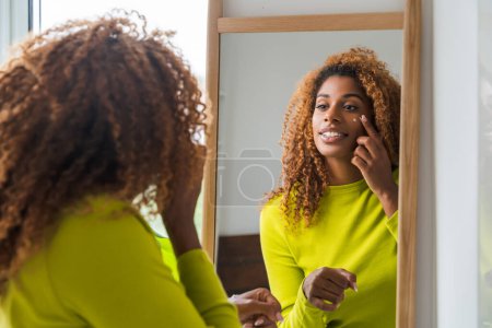 Photo for Back view of the african american beautiful woman standing in front of the mirror and applying cream on face. People lifestyle concept - Royalty Free Image