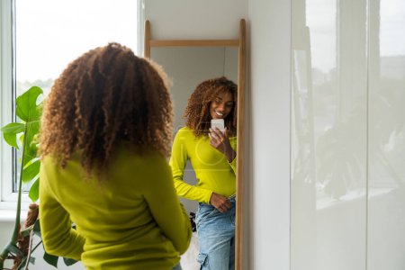 Photo for African american beautiful woman standing in front of the mirror and making smartphone photo of her reflection. People lifestyle concept - Royalty Free Image