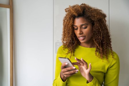 Photo for Curly african american woman holding mobile phone and typing message while standing at home. Technologies and people concept - Royalty Free Image