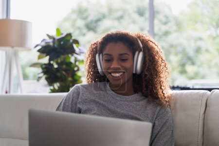 Photo for Attractive african american woman is using a computer and listening music in headphones at living room. Technologies and people concept - Royalty Free Image