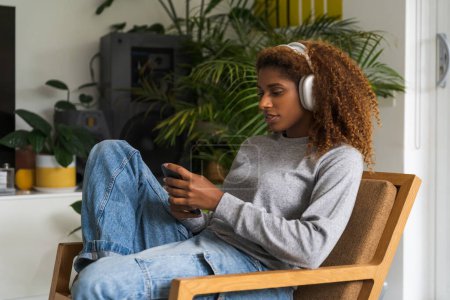 Photo for Curly girl wearing headphones sitting at the sofa and looking at the mobile phone screen. People and technologies concept - Royalty Free Image