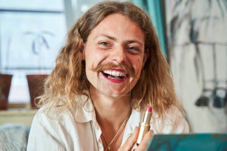 Photo for Portrait of the laughing longhaired guy with moustaches holding pink lipstick and looking at camera at home. People lifestyle concept - Royalty Free Image