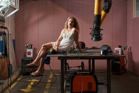 Photo for Feminine energetic carpenter is standing at his job. Longhaired guy in lady dress sitting at the table while posing at workplace - Royalty Free Image