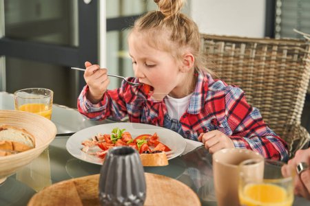 Photo for Focused child girl holding fork and eating with appetite while enjoying healthy breakfast at light kitchen. Family concept - Royalty Free Image