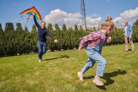 Photo for Little blonde girl running at summer green meadow while playing with father. Happy man holding rainbow kite at background - Royalty Free Image