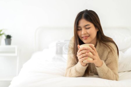 Téléchargez les photos : Portrait de sourire heureuse beauté gaie jolie femme asiatique détente boire et regarder tasse de café chaud ou thé.Girl abattage profiter d'avoir le petit déjeuner en vacances matin vacances sur le lit à la maison - en image libre de droit