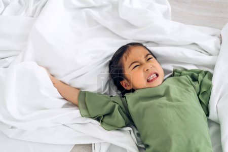 Téléchargez les photos : Portrait de heureux souriant petit enfant asiatique fille s'amuser couché dans le lit blanc et en regardant la caméra à la maison - en image libre de droit