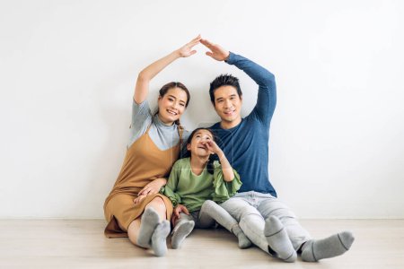 Téléchargez les photos : Portrait profiter heureux sourire amour asiatique famille père et mère avec les jeunes parents petite fille asiatique assis et faire maison sur le toit avec les mains bras sur la tête dans la nouvelle maison - en image libre de droit