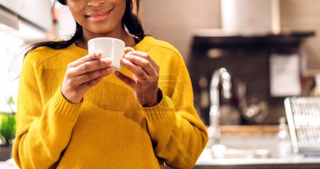 Téléchargez les photos : Portrait de souriante femme noire afro-américaine heureuse relaxante buvant et regardant une tasse de café ou de thé chaud.Fille abattage profiter de prendre le petit déjeuner en vacances matin vacances à la maison - en image libre de droit