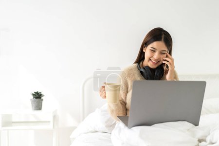 Téléchargez les photos : Jeune souriant heureux belle asiatique femme travail relaxant en utilisant ordinateur portable sur canapé à la maison.Jeune fille créative travaillant et utiliser smartphone.work concept de la maison - en image libre de droit