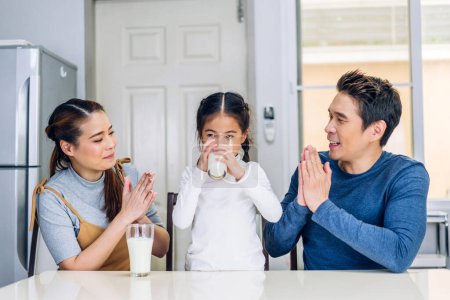 Téléchargez les photos : Portrait de jouir heureux amour asiatique famille père et mère avec petite fille asiatique souriant et ayant protéines petit déjeuner boire et tenir des verres de lait à table dans la cuisine - en image libre de droit