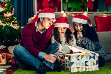 Téléchargez les photos : Portrait de père et mère heureux de famille avec fille dans chapeaux de père Noël avoir plaisir à ouvrir boîte cadeau de Noël magique et profiter de passer du temps ensemble dans le temps de Noël à la maison - en image libre de droit