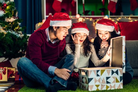 Téléchargez les photos : Portrait de père et mère heureux de famille avec fille dans chapeaux de père Noël avoir plaisir à ouvrir boîte cadeau de Noël magique et profiter de passer du temps ensemble dans le temps de Noël à la maison - en image libre de droit