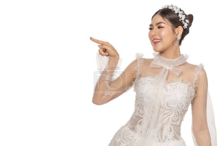 Téléchargez les photos : Portrait de sourire joyeux belle femme mariée de mode asiatique posant en robe de mariée blanche et pointant vers le haut espace de photopour la publicité sur fond blanc - en image libre de droit