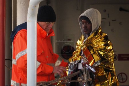 Foto de Migrante sale del barco acompañado por un voluntario de protección civil. El barco de rescate alemán "Sea-Eye 4" llega al puerto de Nápoles con 109 migrantes a bordo, incluidos 35 menores de edad, 18 mujeres, una de las cuales está embarazada y dos fallecidas.. - Imagen libre de derechos