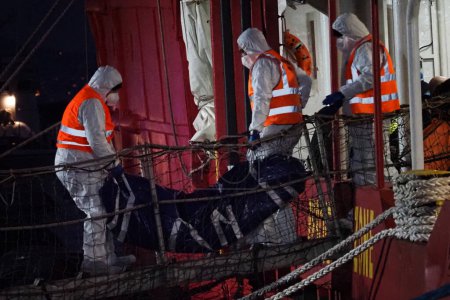 Foto de Oficiales de policía mortuorios sacan el cuerpo de un migrante de la nave. El barco de rescate alemán "Sea-Eye 4" llega al puerto de Nápoles con 109 migrantes a bordo, incluidos 35 menores de edad, 18 mujeres, una de las cuales está embarazada y dos fallecidas.. - Imagen libre de derechos