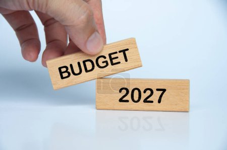 Mano sosteniendo bloque de madera con Presupuesto 2027 texto sobre fondo blanco. Concepto de presupuestación anual