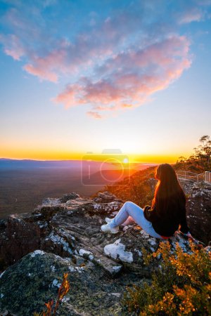 Frau sitzt am Rand der Reed Lookout-Klippe und genießt den Sonnenuntergang über dem Victoria Valley in den Grampians Mountains, Victoria, Australien