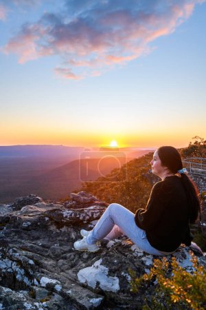 Femme assise sur le bord de la falaise Reed Lookout tout en profitant du coucher de soleil au-dessus de la vallée de Victoria dans les montagnes des Grampians, Victoria, Australie