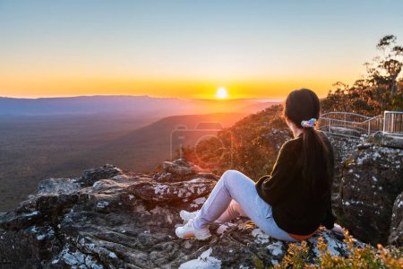 Junge Frau sitzt am Rande der Klippe des Reed Lookout und genießt den Sonnenuntergang in den Grampians Mountains, Victoria, Australien