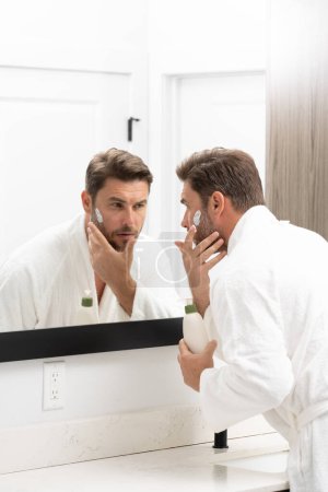 Young happy manful guy applying face cream. Close up shot of attractive man with healthy skin, applies cream for anti wrinkle or anti aging, cares of body. Beauty portrait. Body positive. Skin