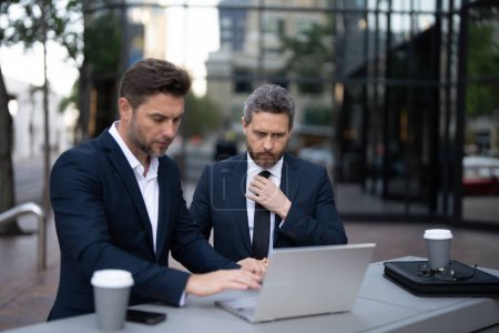 Zwei Geschäftsleute auf der Straße, tief verstrickt, die Augen starr auf ihren Laptop gerichtet, diskutieren vielleicht einen entscheidenden Deal oder planen ihr nächstes großes Projekt im geschäftigen Stadtbild..